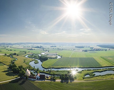 Landschaft mit einem sich schlängelndem Fluss, Feldern und Gebäuden, unter einem klaren Himmel mit strahlender Sonne.