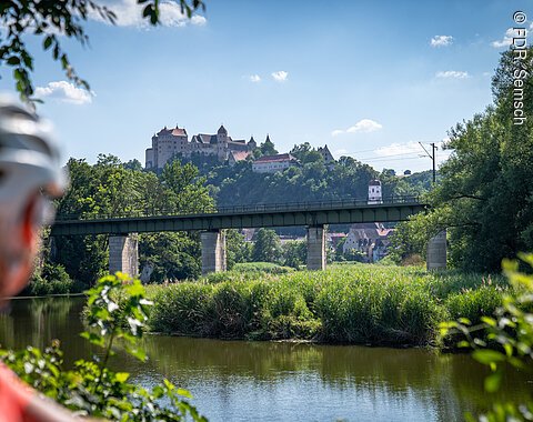 Blick auf die Eisenbahnbrücke über der Wörnitz mit der Harburg im Hintergrund, Fahrradhelm einer Radfahrerin im Vordergrund.