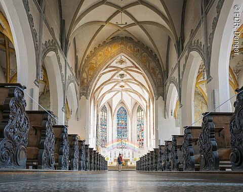 Innenansicht einer Kirche mit verzierten Holzbänken, gotischen Bögen und einem bunten Glasfenster im Hintergrund.