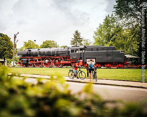 Radler vor der Eisenbahn in Treuchtlingen Dampflokomotive auf Gleis mit zwei Radfahrern davor, umgeben von Bäumen und Wiese bei bewölktem Himmel.