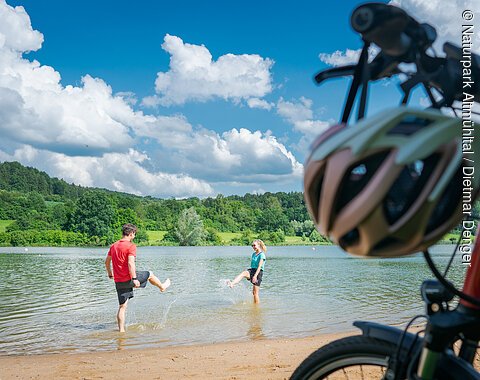Am Hahnenkammsee Im Vordergrund stehen rechts zwei Fahrräder, dahinter liegt ein kurzer Strand an einem Seeufer. Im seichten Wasser stehen ein Mann und in eine Frau in kurzen Hosen und T-Shirts, die versuchen, sich gegenseitig nass zu spritzen. Am Ufer gegenüber liegen eine Wiese und Wald.