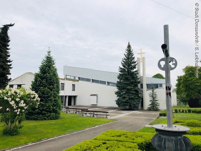 Blick auf die Kirche. Das Gotteshaus ist weiß, ein moderner Turm mit Kreuz, außen sind Bäume, Sträucher usw.