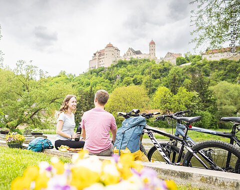 Zwei Personen sitzen auf einer Wiese mit Fahrrädern, im Hintergrund die Harburg auf einem Hügel.