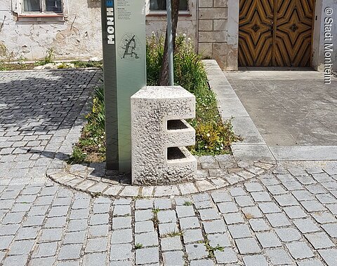 Buchstabenweg Monheim Steinskulptur des Buchstabens "E", dahinter ein Informationsschild und ein Baum mit einem dahinter liegendem Beet. Umgeben von einem gepflastertem Platz, mit einem historischen Gebäude im Hintergrund.