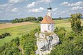 Kleine Kirche mit Zwiebelturm auf einem Felsen, umgeben von grünen Feldern und Bäumen.
