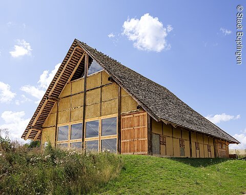 Blick auf ein Gebäude des Freilichtmuseums am Ipf Großes Fachwerkhaus mit steilem Schindeldach auf einer grünen Wiese unter blauem Himmel mit Wolken.