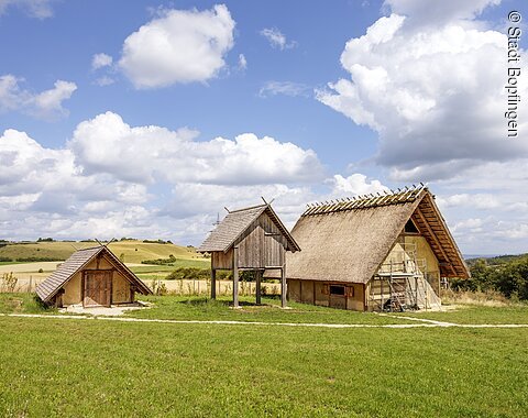 Blick auf rekonstruierte Gebäude am Ipf Drei historische Gebäude mit Strohdächern auf einer Wiese unter blauem Himmel mit Wolken.