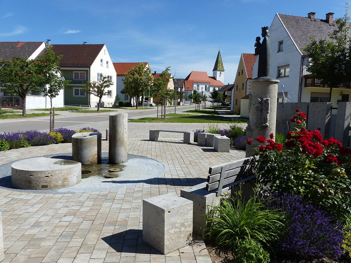 Blick auf den Nadlerbrunnen mit Sitzgelegenheiten. Im Hintergrund sieht man die Kirche.