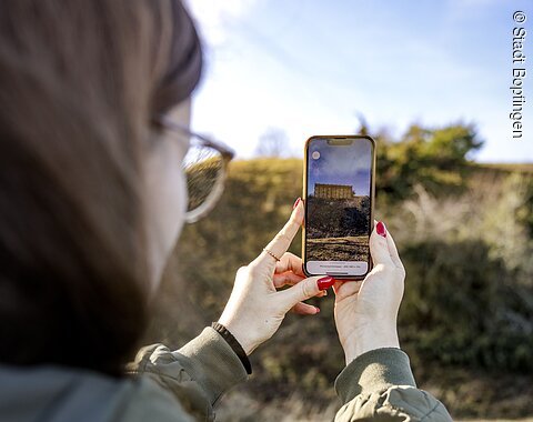 Digitaler Rundgang am Ipf Frau mit Brille und rotem Nagellack hält Smartphone mit Bild einer historischen Pfostenschlitzmauer im Freien.