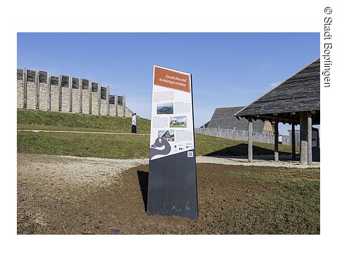 Infotafel am keltischen Freilichtmuseum mit Beschreibungstext und QR-Code zum digitalen Rundgang am Ipf Informationstafel mit dem Titel „Geschichte und Archäologie erleben“ vor einer historischen Holz- und Steinmauer bei klarem Himmel.