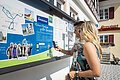 Startpunkt der Lauschtour "Kleines Monheim, große Geschichte"- an der Tourist-Information Monheim Zwei Personen vor einem weißen Gebäude, der Touristinformation Monheim betrachten eine Informationstafel mit einer Karte und Texten zur Lauschtour in Monheim und haben ein Smartphone in der Hand, mit dem sie einen Code abscannen.