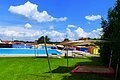 Freibad Monheim - Sandkasten mit Sonnensegel Ein Freibad mit Schwimmbecken, Sonnensegeln, Sitzmöglichkeiten und einem Sandkasten auf einer grünen Wiese, rechts unten im Bild. Im Hintergrund blauer Himmel mit Wolken.