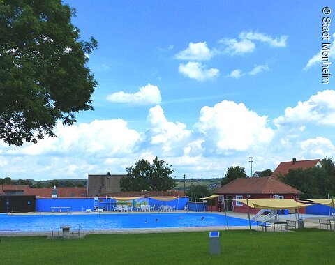 Freibad Monheim "Nostalgiebad" Freibad mit blauem Wasser, umgeben von Rasen, Bäumen und Gebäuden unter einem bewölkten Himmel. Vorderhalb vom Becken sind Sitzmöglichkeiten.