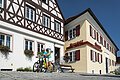 Tourist-Information der Stadt Monheim/Monheimer Alb - im Schindlerhaus Tourist-Information der Stadt Monheim/Monheimer Alb: ein Fachwerkhaus mit drei Stockwerken, weißen Wänden und braunen Balken. Rechts davon ein gelbes zweistöckiges Gebäude mit roten Fensterläden. Zwei Personen mit Fahrrädern stehen davor und schauen eine Broschüre an.