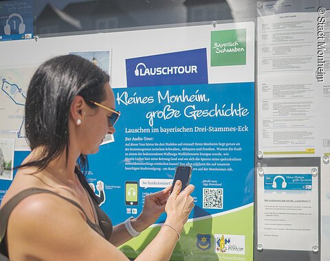 Frau mit Smartphone steht vor Infotafel zur Lauschtour in Monheim, Bayern, mit dem Titel "Kleines Monheim, große Geschichte" bei Sonnenschein. Frau hält ihr Smartphone an einen QR-Code.