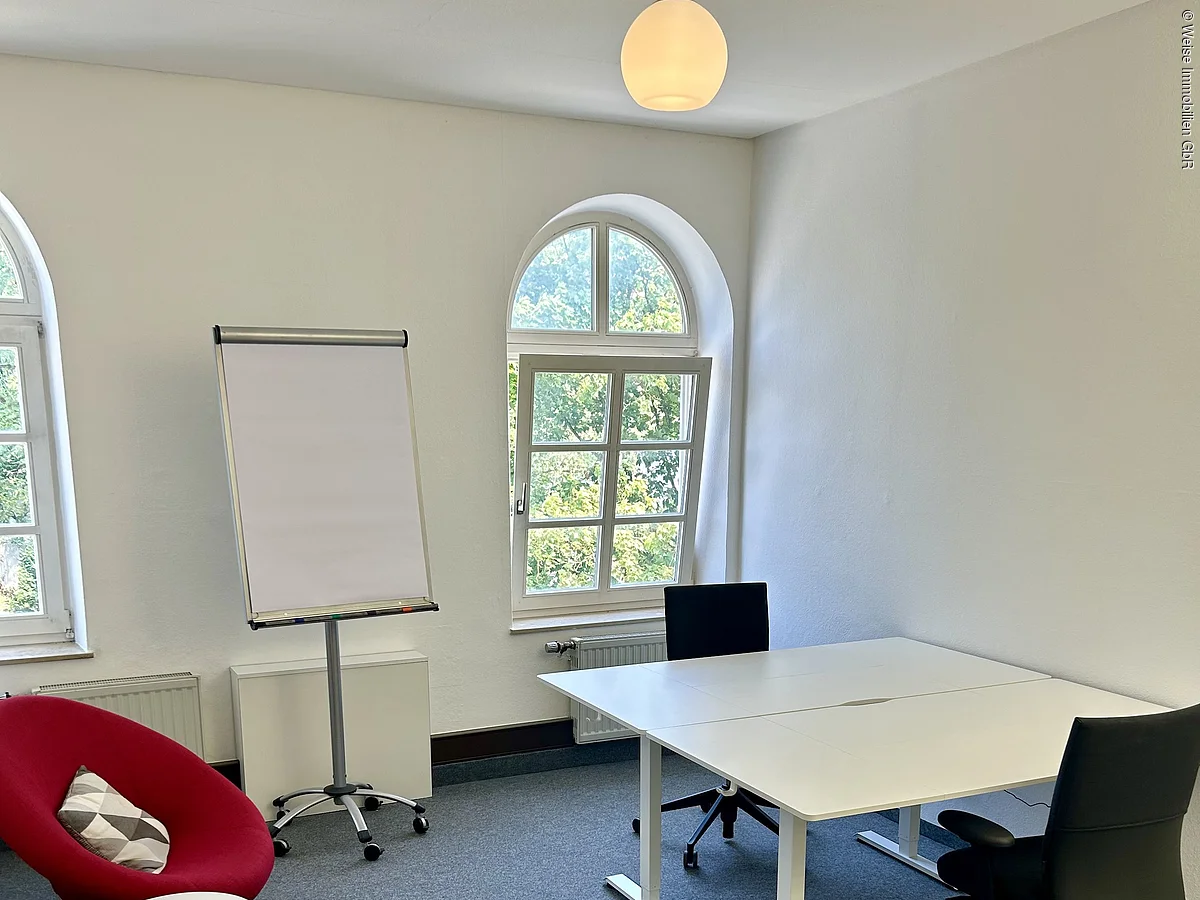 Büro mit zwei weißen Tischen, zwei schwarzen Bürostühlen, Flipchart, zwei Sesseln und zwei Fenstern mit Rundbogen.