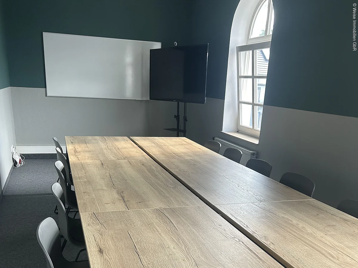 Konferenzraum mit langem Holztisch, Stühlen, großem Fenster, Whiteboard und Bildschirm an der Wand.