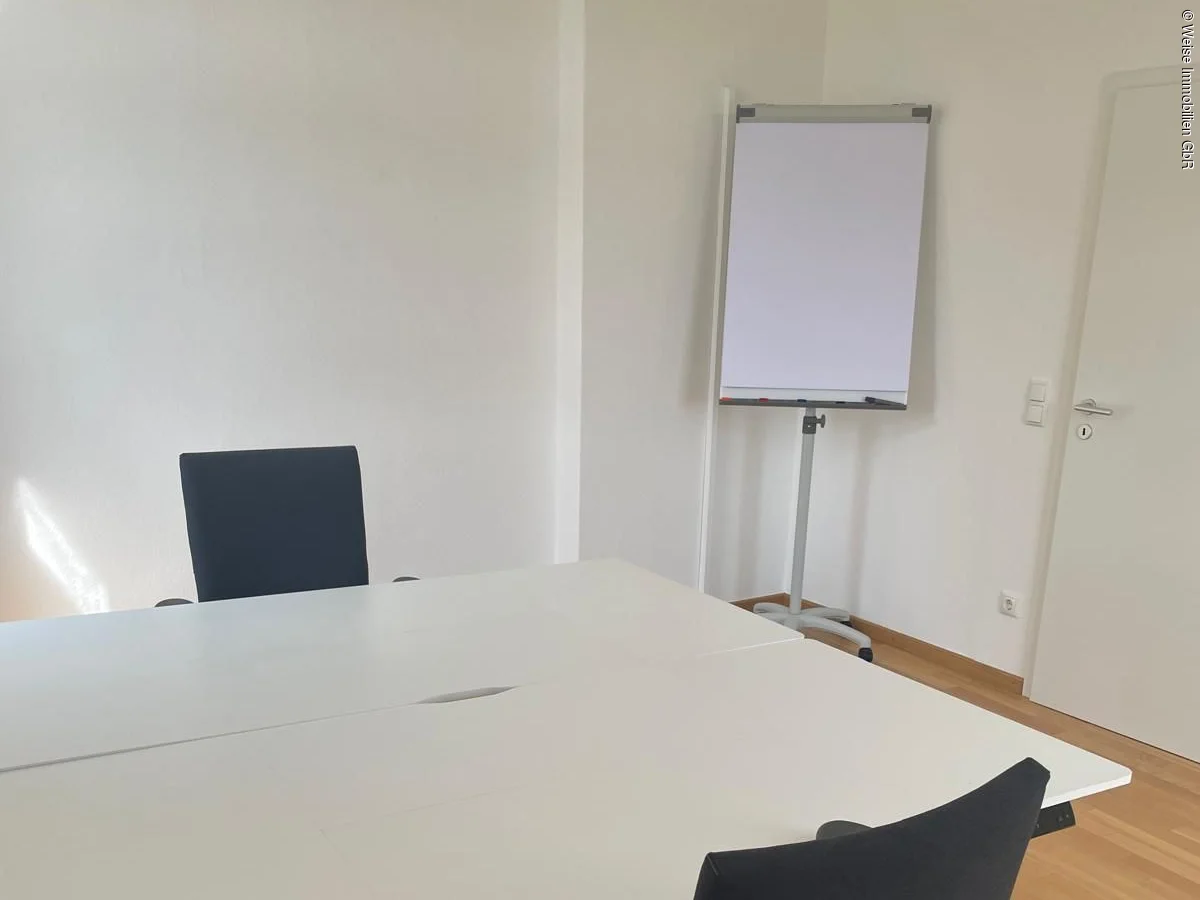 Büro mit zwei schwarzen Bürostühlen, weißen Tischen, Flipchart und Holzboden.