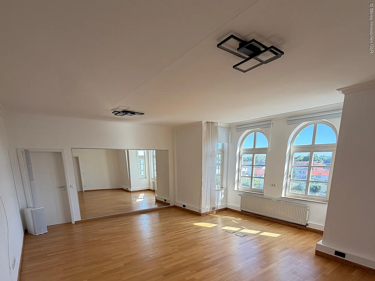 Leerer Raum mit Holzboden, zwei großen Bogenfenstern und einer Wand mit großem Spiegel.
