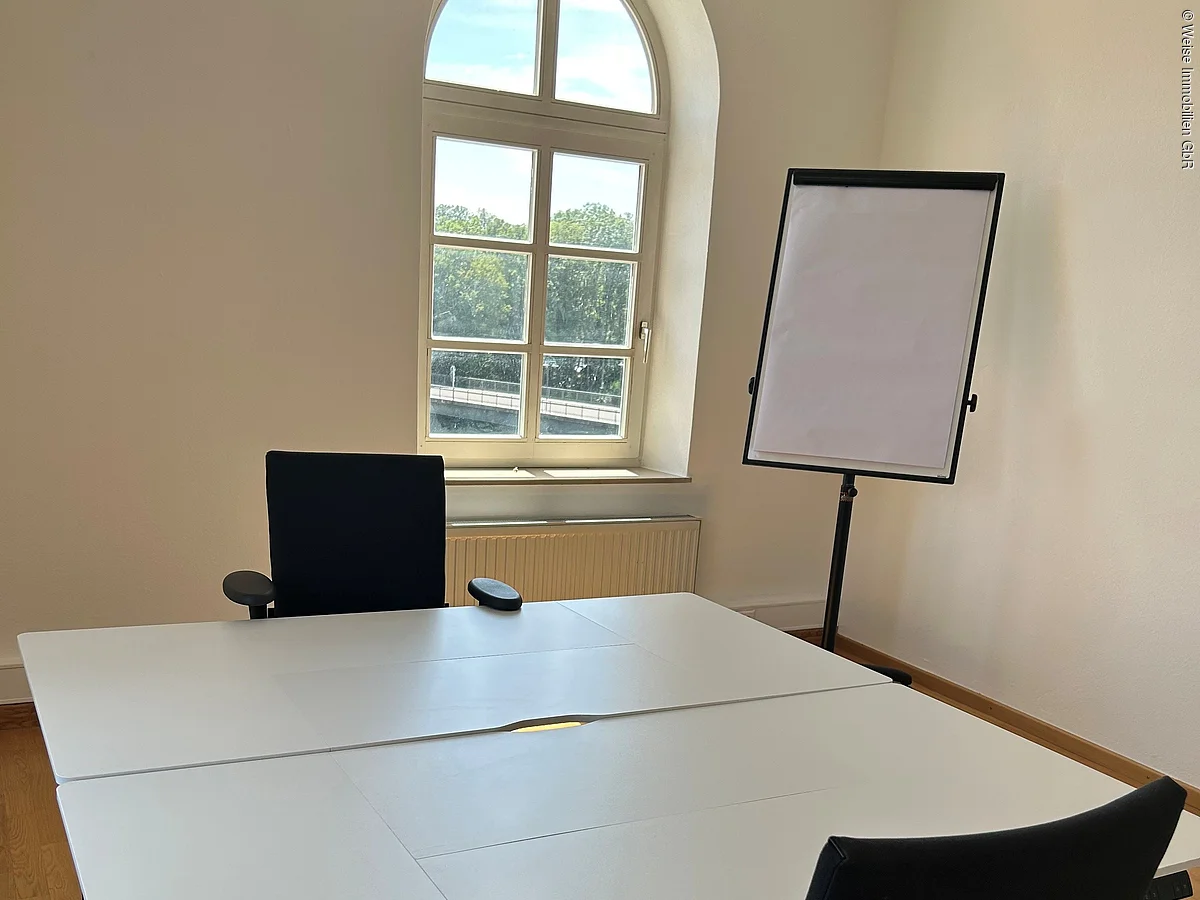 Büro mit zwei schwarzen Bürostühlen, weißem Tisch, Flipchart und rundem Fenster mit Blick nach draußen.