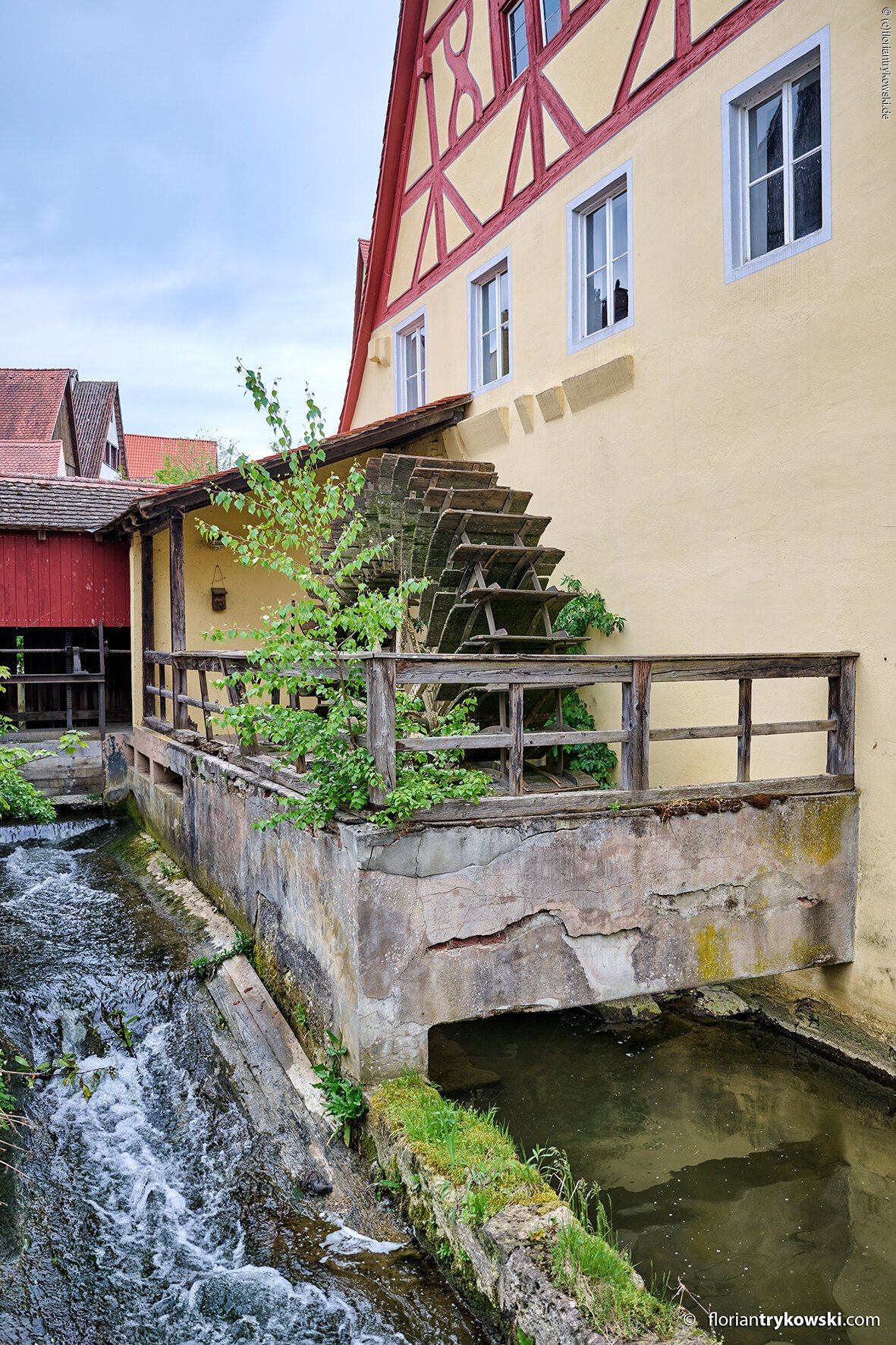 Altes Wasserrad an gelbem Fachwerkhaus neben fließendem Bach mit Holzgeländer und Pflanzen im Nördlinger Gerberviertel
