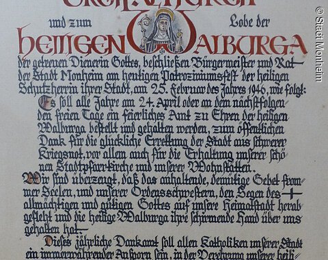 Dankamt-Urkunde von 1946 von Bürgermeister und Rat der Stadt Monheim Ein handgeschriebenes Dokument mit dekorativen roten und schwarzen Buchstaben, das eine religiöse Widmung enthält. Hierin gelobt der Bürgermeister sowie der Rat der Stadt Monheim, ein alljährliches Dankamt zu Ehren der Heiligen Walburga abhalten zu lassen, für die Verschonung der Stadt im Zweiten Weltkrieg.