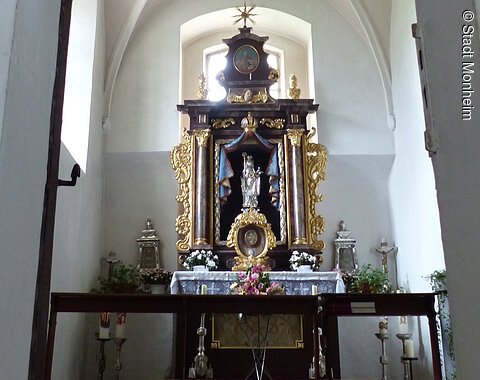 Walburgakapelle in der Stadtpfarrkirche St. Walburga Monheim Ein Blick durch eine Tür zu einem Kirchenaltar in der Walburgakapelle in Monheim, mit goldenen Verzierungen und einer Statue, der heiligen Walburga. Der Kirchenaltar ist umgeben von Kerzen und Blumen, in einem gewölbten Raum mit einer verzierten Decke.