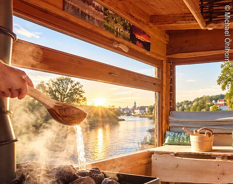 Eine Hand gießt Wasser auf heiße Steine in einer Sauna mit Fluss- und Sonnenuntergangsblick durchs Fenster.